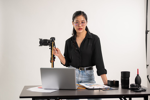 An attractive, professional Asian female photographer producer stands at a desk in a photoshoot studio with professional photography equipment on the desk.
