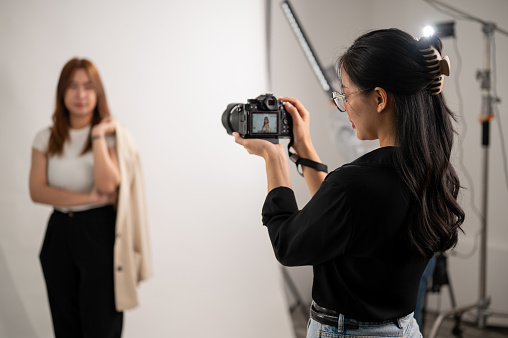 A professional, cool Asian female photographer taking pictures of a model in the studio with professional lighting equipment. A team of fashion photoshoot working in the studio.