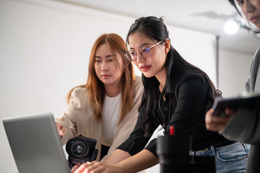 A professional Asian female photographer is working with her assistants in a photoshoot studio, talking, briefing the concept, checking images on the laptop, and working as a team.