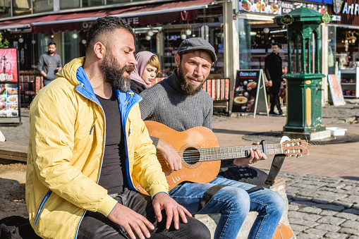 Istanbul, Turkey - December 29, 2022: Two men are sitting on bench by the street and one of them playing guitar.