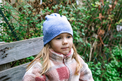 A cute preschool girl with long blond hair and blue eyes sitting on a wooden bench in the garden wearing a red and white fleece jacket and blue beanie, looking happy at the camera.