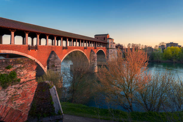 amazing view of Ponte Coperto Pavia (covered bridge) at sunset wonderful view of Ponte Coperto (covered bridge) is a bridge over the Ticino river in Pavia at sunset, Lombardy, Pavia, Italy clear sky night sunset riverbank stock pictures, royalty-free photos & images