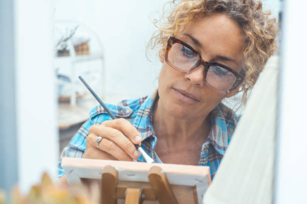 istock Concept of art, creativity, hobbies, work and creative occupation. Front view of blond, curly woman sitting indoors at home in front of a window in front of easel, painting with brush and colors 2114118523