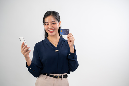 An attractive Asian woman is holding a smartphone and showing a credit card to the camera while standing against a white studio background. promoting online payment and cashless transactions.