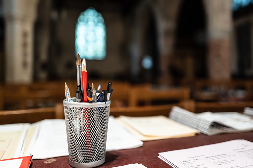 Shallow focus of s selection of pens and pencils seen in an English church next to printed church materials. A large stained glass window can be seen in the alter section.