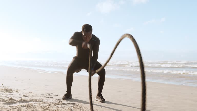 Beach, battle rope and black man with exercise, energy and workout with strength, power and endurance. Seaside, African person and bodybuilder with ropes, training and ocean with cardio and fitness