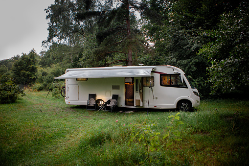 tranquil scene in the heart of nature, where a motorhome is parked in a forest clearing. Chairs are set up outside, and the awning is extended, creating a cozy and inviting atmosphere.