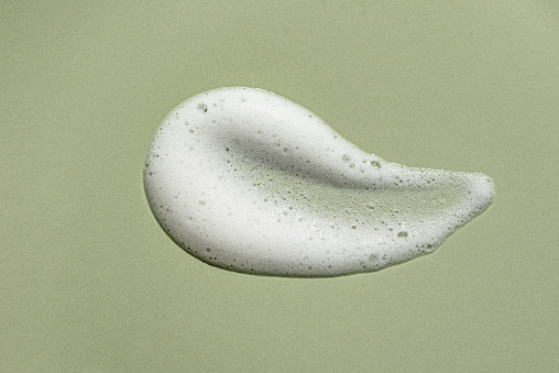 Face cleansing mousse sample. White cleanser or soap foam bubbles on green background. Shower gel, hair shampoo foam texture closeup.