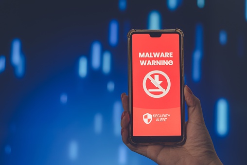 Hand holding a smartphone displaying a malware warning against a blue bokeh background, suggesting themes of cybersecurity and online safety.