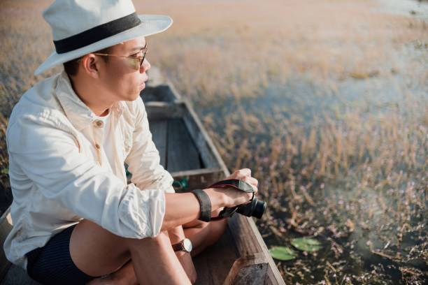 A man in a hat sits in a boat holding a camera, surrounded by calm waters and nature, reflecting a serene, exploration-themed setting.