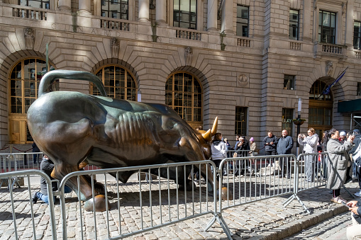 Wall Street, Manhattan, New York, USA - March, 2024.  Tourists getting photographed beside the Bull statue (created by Italian artist Arturo Di Modica in the wake of the 1987 Black Monday stock market crash) in Wall Street, the financial district in New York.  The statue is fenced off to prevent crowds gathering and blocking the road.