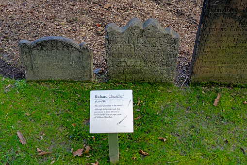 A very old, moss covered, and cracked grave marker in a cemetery. No marking visible. Other tombstones behind. Broken at top.