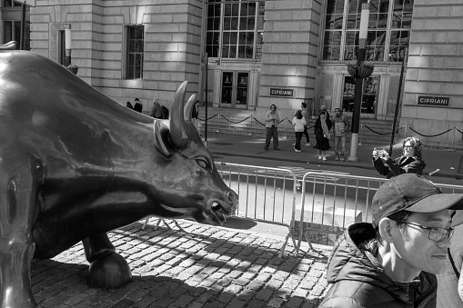 Wall Street, Manhattan, New York, USA - March, 2024.  Tourists getting photographed beside the Bull statue (created by Italian artist Arturo Di Modica in the wake of the 1987 Black Monday stock market crash) in Wall Street, the financial district in New York.  The statue is fenced off to prevent crowds gathering and blocking the road.