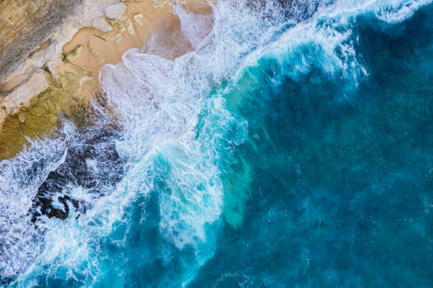 Bali - Aerial view of ocean waves crashing against a rugged shoreline, highlighting the contrast between the deep blue sea and beige rocks. The turquoise water churns with white foam, creating intricate textures visible from above. The jagged rocks, weathered by the relentless waves, form pools of water in various spots.