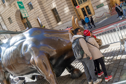 Wall Street, Manhattan, New York, USA - March, 2024.  Tourists getting photographed beside the Bull statue (created by Italian artist Arturo Di Modica in the wake of the 1987 Black Monday stock market crash) in Wall Street, the financial district in New York.  The statue is fenced off to prevent crowds gathering and blocking the road.