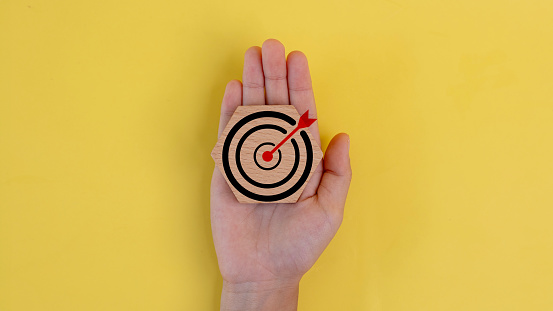 A hand holding a wooden target with a red arrow pointing to the center. Concept of aiming for a goal or target, and the importance of focus and determination in achieving it