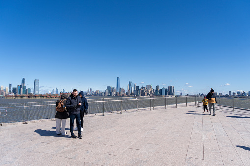 Liberty Island, New York, USA - March, 2024.  Liberty island with tourists in the foreground near the flag pole in front of the Statue of Liberty museum. In the background are the skyscrapers of Wall Street and Lower Manhattan and some tourists are having their photograph taken on a mobile phone with the Manhattan skyline in the background.