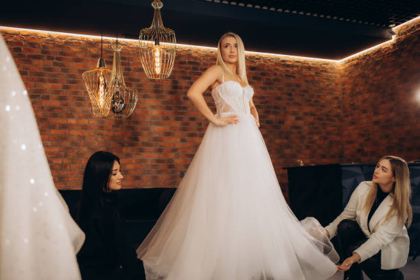 istock Beautiful bride getting dressed by her best friend in her wedding day and choosing a wedding dress in the shop and the shop assistant is helping her 2114341575