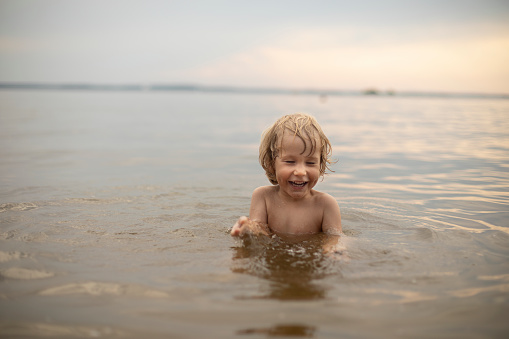 A three-year-old fair-haired boy bathes outdoors on a summer evening in the water, Holidays with children, restoration and relaxation in nature, development of children's scientific and cognitive activity in the open air, filling with joy and emotions, happy childhood and restoration of mental health