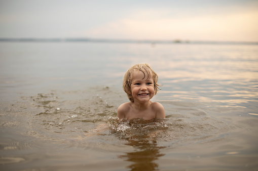 A three-year-old fair-haired boy bathes outdoors on a summer evening in the water, Holidays with children, restoration and relaxation in nature, development of children's scientific and cognitive activity in the open air, filling with joy and emotions, happy childhood and restoration of mental health