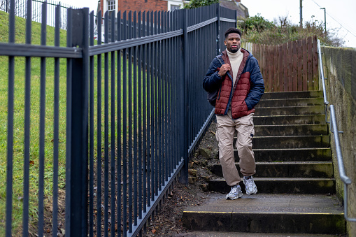Full length wide front view of a young man walking to work on a cold morning in Newcastle, England. He is wearing warm clothing, carrying a backpack. He is walking down city steps.