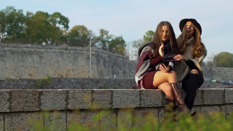 Two Young Elegant Women Using A Phone Together While Sitting On The Wall On The Quayside