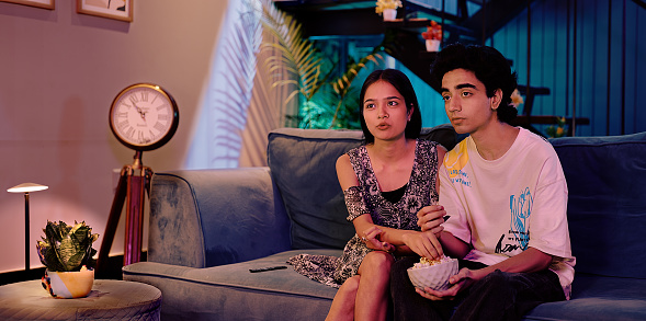 Two terrified Indian boyfriend with girl sitting on sofa watching horror film TV at home late night spend time together. Beautiful teen friend couple excited eat popcorn snack looking action video