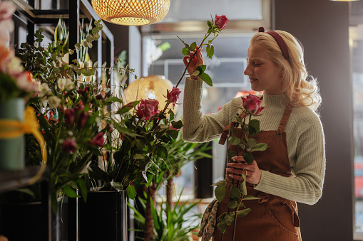 A blonde woman in a brown apron holds and examines roses delicately at a flowering commerce store, with lighting fixtures in the background
