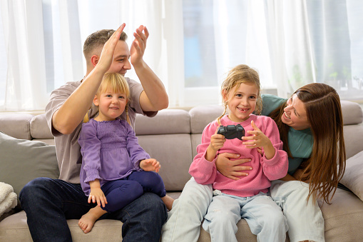 Parents celebrating daughter play video game while sitting on sofa in living room at home.