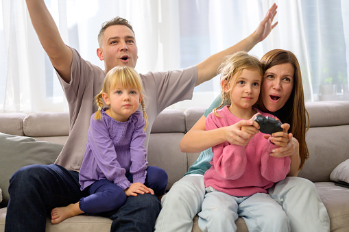 Parents watching daughter play video game while sitting on sofa in living room at home.