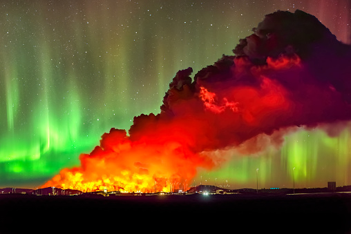 Eruption of the volcano near Grindavik in 2024 seen from the City of Keflavík at night with glowing Northern Lights in the sky