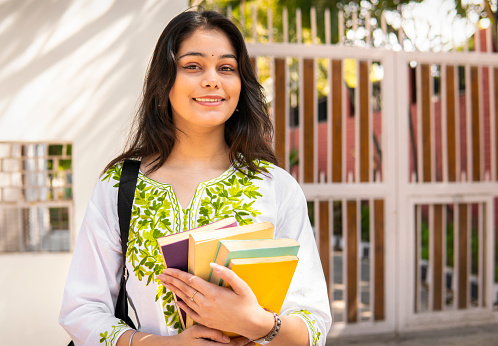 Outdoor portrait of happy beautiful indian young female university student holding books in hand and looking at the camera with confident toothy smile.