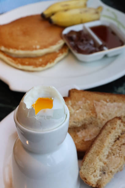 Stock photo showing close-up view of white crockery of tea plate and egg cup with white toast soldiers and topped soft-boiled egg showing runny yellow yolk.