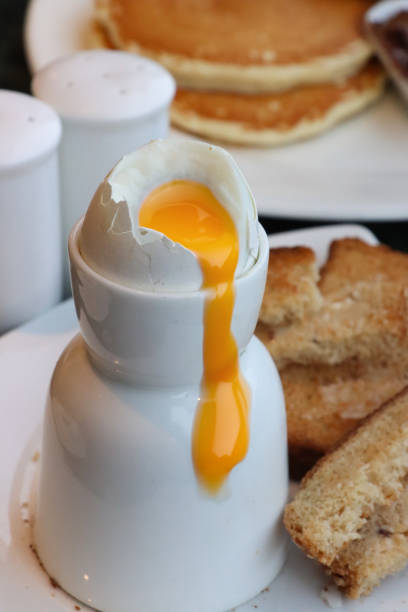 Close-up image of topped soft boiled egg with runny yolk in white egg cup with white buttered toasted bread slices on white plate, salt and pepper shakers, plate containing two, fluffy, American pancakes served with dish of chocolate spread