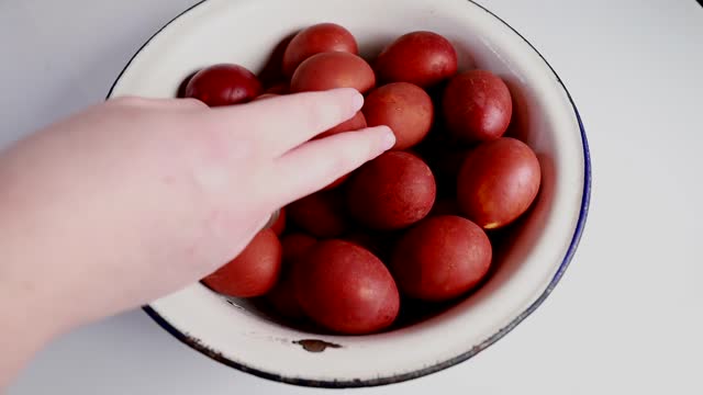 A large bowl filled with Easter eggs is on a table. A woman's hand reaches out to take a red egg
