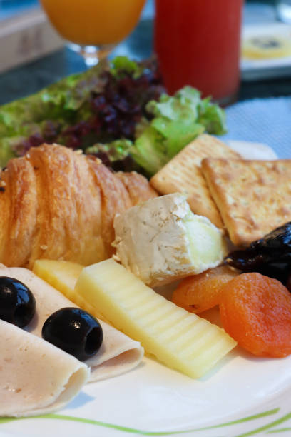 istock Close-up image of white plate containing hotel self service breakfast buffet ingredients, self service continental breakfast, croissant, dry crackers, brie, cheddar cheese, chicken ham, dried apricots, black olives, lettuce leaves, glasses of fruit juice 2114673491