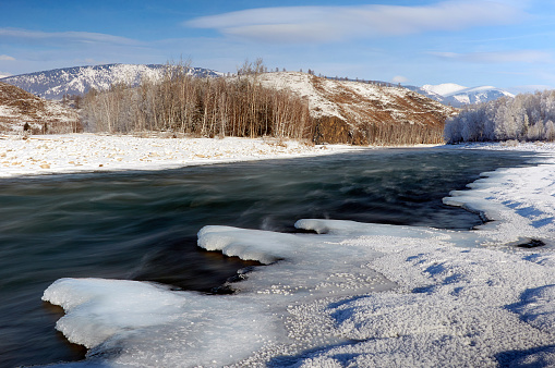 A river flows with ice along its shore, framed by a majestic mountain in the background. The sky is filled with clouds, creating a beautiful natural landscape. Russia, Chuisky ridge, Katun river.