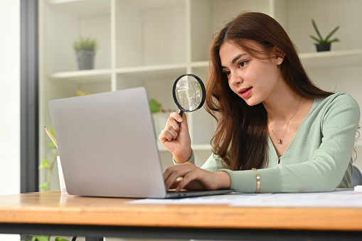 Beautiful young businesswoman hand holding magnifying glass and using laptop computer at office desk.