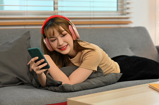 Young Asian girl with headphones listening to music pleasantly on mobile phone as she lying down comfortably on the sofa.