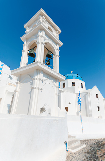 The tranquil beauty of Saint Anastasios Church in Imerovigli on a radiant summer's day. The impressive bell tower rises majestically against the clear blue sky, while the dome, painted in hues of the Aegean Sea, adds to the enchanting charm of this historic site. Whitewashed walls bathe in sunlight, casting a warm glow over the surroundings and inviting visitors to bask in the serenity of this sacred space. With its harmonious blend of architecture and natural beauty, Saint Anastasios Church stands as a timeless symbol of peace and tranquility in the heart of Thira, Santorini.
