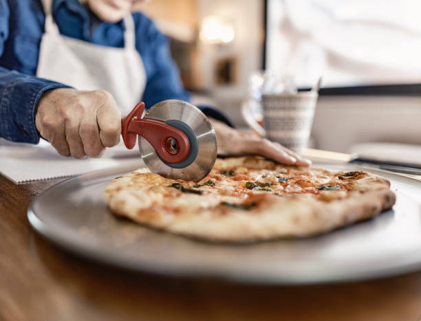 Close up of hand of Senior man cutting pizza while enjoying RV trip adventure. He is dressed in casual clothing. Interior of  recreational vehicle during the day in Ontario, Canada.