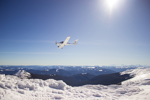Small white airplane flying over mountains and mountain ranges against sunlight. The plane is clearly visible and completely falls into the frame