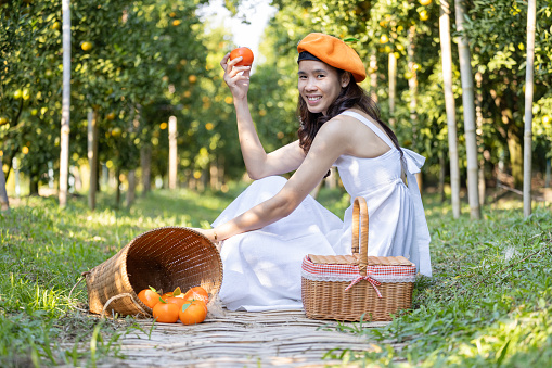 Smiling woman wearing a playful orange-shaped hat holds a fresh orange fruit in a sunny, green garden setting.