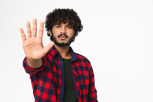 Serious young Hindi man showing stop gesture with palm isolated over white background. Indian handsome boy expressing dismissal gesture, forbidden concept