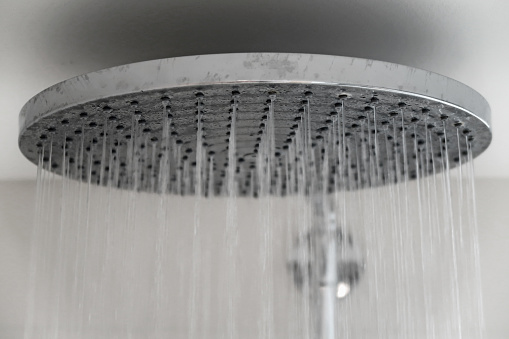 Multiple jets of streaming water pouring from a large, round, silver coloured rain shower head. Washing and personal hygiene. Close up interior design detail.
