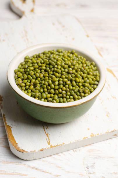 Green mung beans in a ceramic bowl on a white cutting board. Organic legumes. Vegan and vegetarian food. The concept of healthy eating. Vertical orientation. Selective focus. stock photo