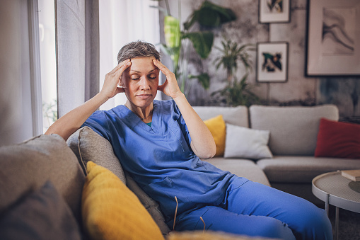 Woman in medical scrubs has a headache while sitting on sofa after work at home.