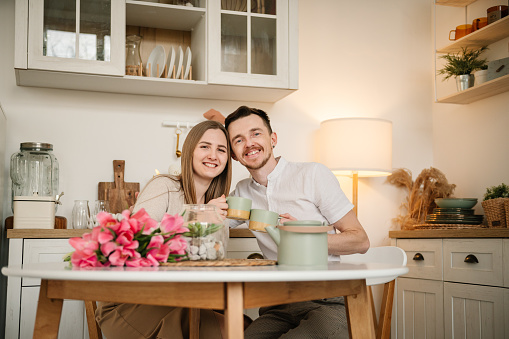Romantic female and male talking spending time together at home. Loving young man and woman having conversation and drink tea in morning. Couple drinking coffee, holding cups in hands in the kitchen.