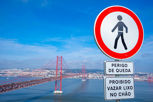 Road sign prohibiting pedestrian movement and danger of falling caption with the background of the metal structure platform of the 25 de Abril bridge from the city of Almada.