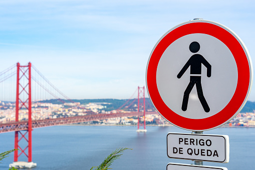 Road sign prohibiting pedestrian movement and danger of falling caption with the background of the metal structure platform of the 25 de Abril bridge from the city of Almada.